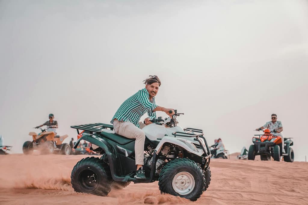 A group of people ride variously colored ATVs across a sandy desert, kicking up dust under a bright sky.
