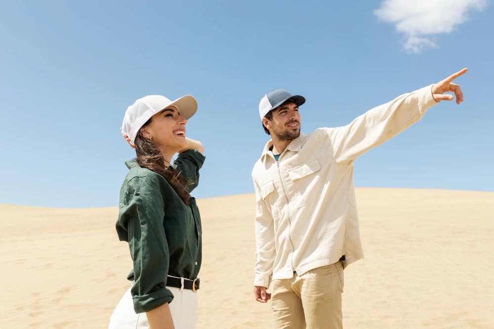 A man and woman wearing light shirts, neutral trousers, and caps in the desert, showcasing practical and sun-protective outfits for what to wear in Dubai desert safari.