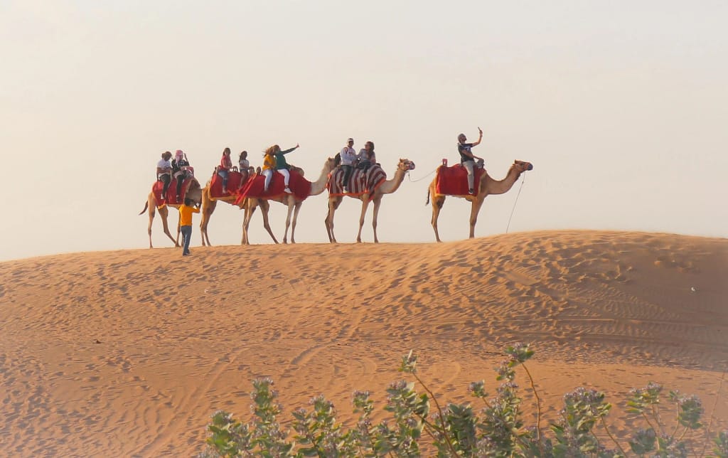 Tourists riding camels on sand dunes with a guide, enjoying a desert safari under a clear sky.