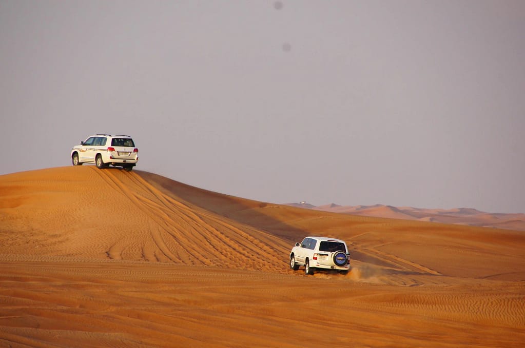 Two white 4x4 vehicles driving across sand dunes in the desert during daylight.
