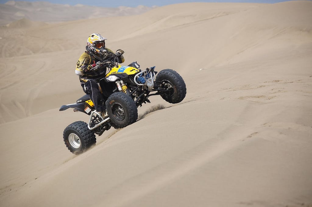A rider wearing protective gear drives a yellow and black ATV up a sandy desert dune, with sand spraying from the tires.