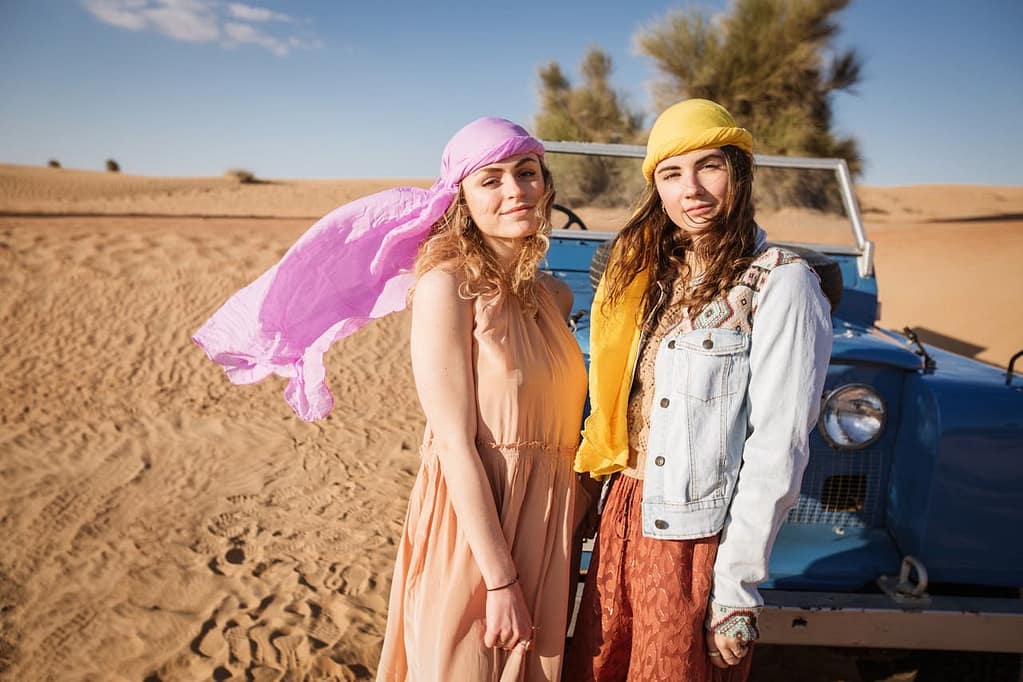 Two women posing near a blue jeep in the Dubai desert, one in a yellow headscarf and denim jacket, the other in a peach maxi dress with a pink scarf, perfectly illustrating what to wear in Dubai desert safari for a fashionable and comfortable look.
