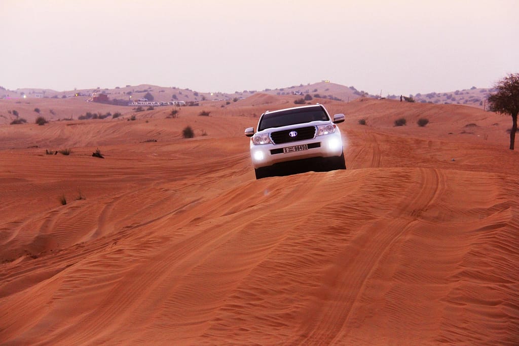 White SUV climbing a sand dune at sunset in a wide desert valley.