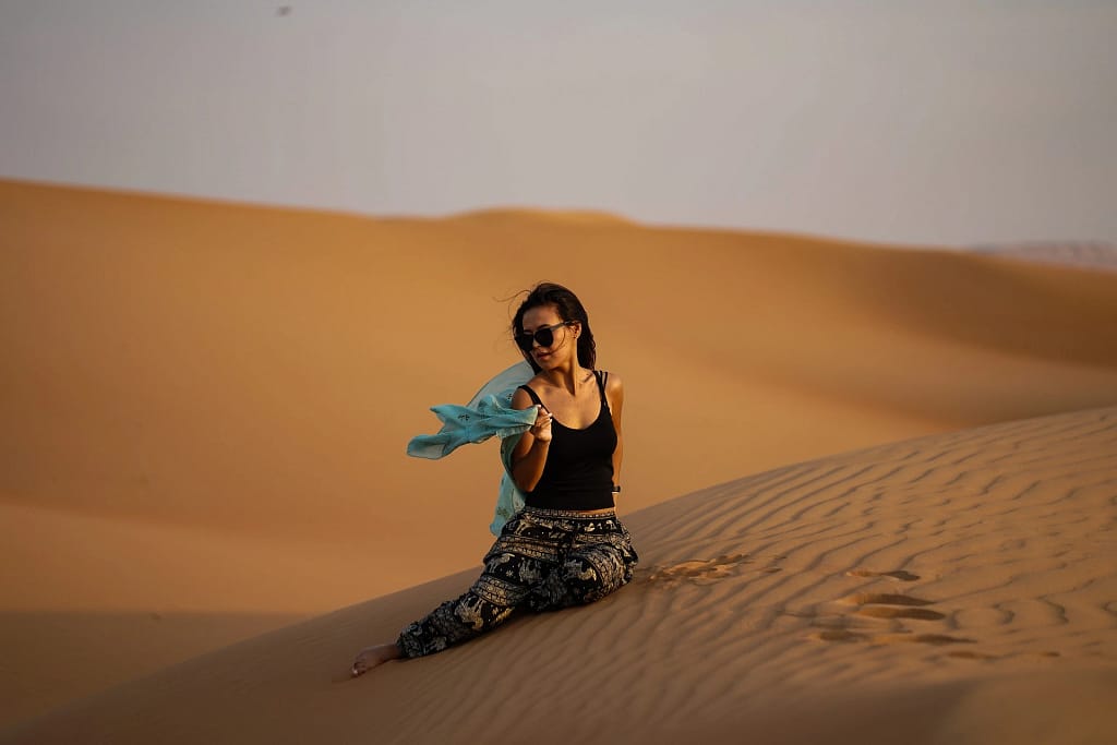 Woman sitting on a sand dune with sunglasses and scarf in hand during golden hour.