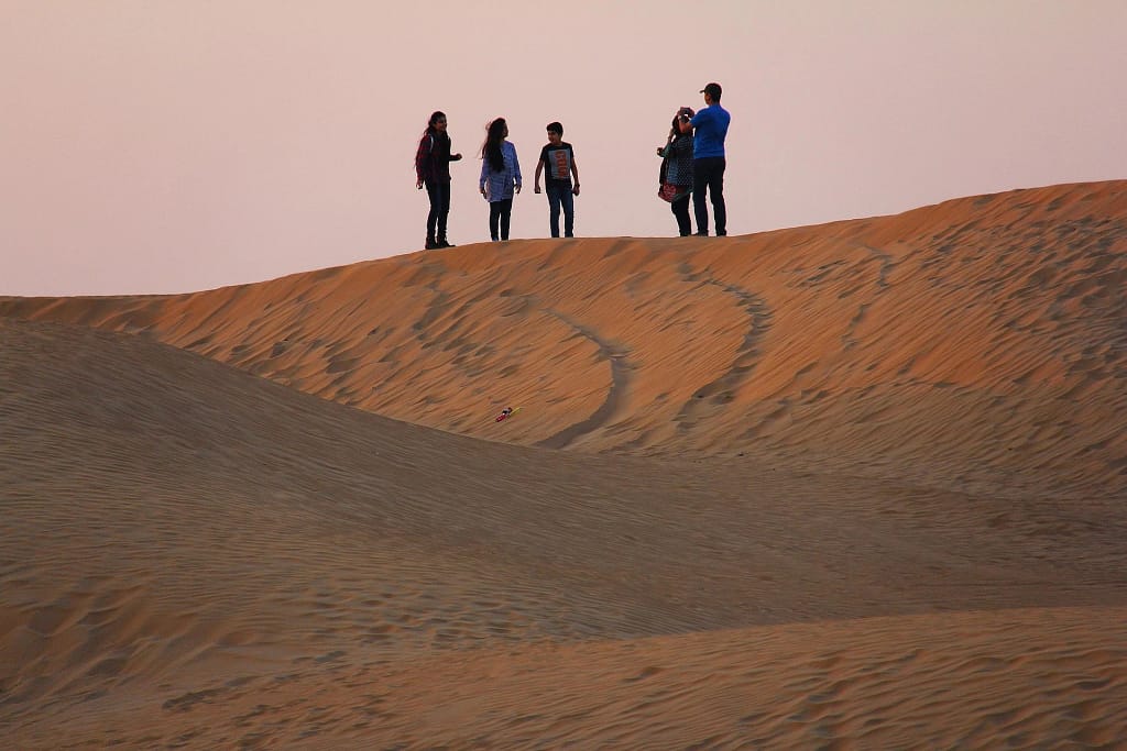 Group of people standing on a sand dune taking photos during a desert sunset.