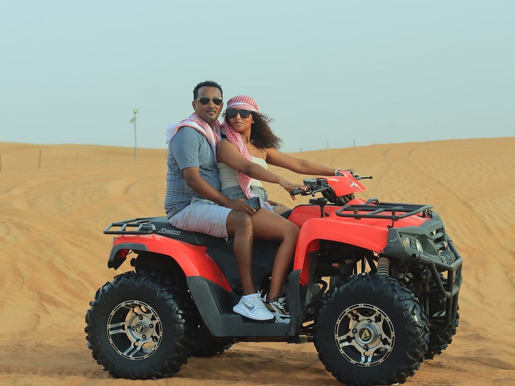 A man and a woman ride a red four-wheeler (ATV) over sand dunes in a vast desert under a clear sky.