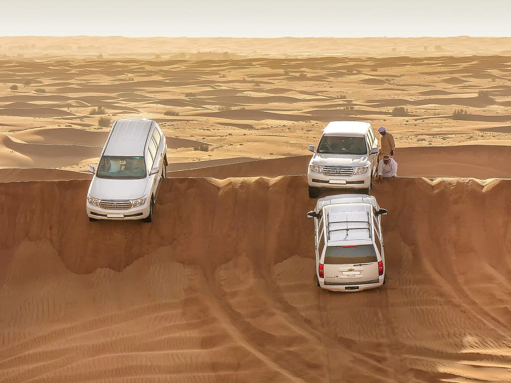 Three white SUVs lined up on the edge of a steep desert dune under bright sunlight.